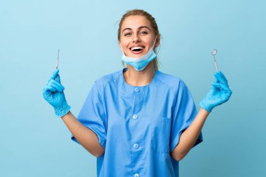 Genç kadın diş hekimi mavi arka planda aletler tutuyor.