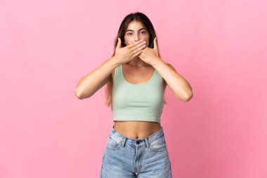 Pembe arka planda izole edilmiş genç beyaz kadın elleriyle ağzını kapatıyor.