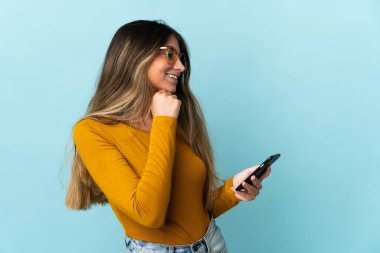 Mavi arka planda izole edilmiş cep telefonu kullanan beyaz bir kadın bir fikir üretiyor ve bakıyor.