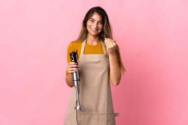 Genç aşçı kadın el blenderi kullanıyor. Pembe arka planda izole edilmiş. Bizimle gelmesi için davet ediliyor. Gelmene sevindim.