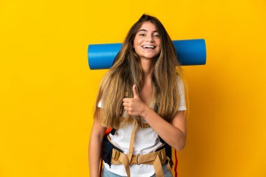 Sarı arka planda izole edilmiş büyük sırt çantalı genç dağcı kadın baş parmak işareti yapıyor.