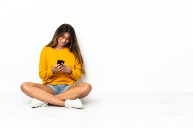 Yerde oturan genç kadın, beyaz arka planda izole bir şekilde cep telefonuyla bir mesaj gönderiyor.