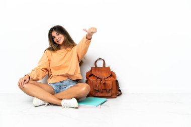 Yerde dizüstü bilgisayarlı genç bir bayan oturuyor ve beyaz arka planda izole edilmiş bir şekilde baş parmak işareti yapıyor.