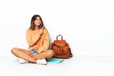 Genç bir bayan öğrenci dizüstü bilgisayarla yerde oturuyor beyaz arka planda izole edilmiş bir şekilde bir ürünü sunmak için kenarı gösteriyor.