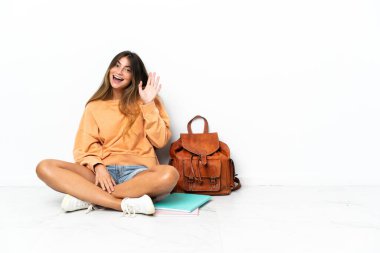 Genç bir kız öğrenci dizüstü bilgisayarla yerde oturuyor. Beyaz arka planda izole edilmiş, mutlu bir ifadeyle selamlıyor.