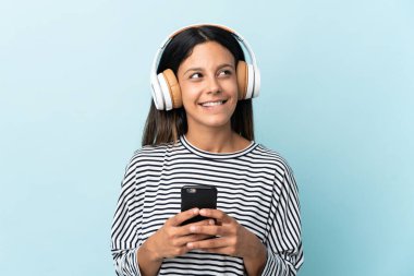 Kafkasyalı bir kız mavi arka planda izole edilmiş. Cep telefonuyla müzik dinliyor ve düşünüyor.