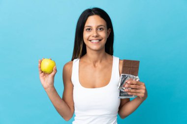 Kafkasyalı kız mavi arka planda izole edilmiş. Bir elinde çikolata tableti, diğer elinde elma var.