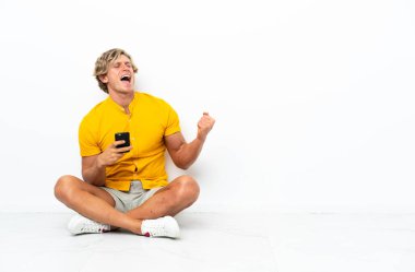 Genç İngiliz adam zafer pozisyonunda telefonla yerde oturuyor.