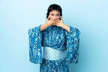 İzole edilmiş mavi arka planda kimono giyen genç bir kadın elleriyle ağzını kapatıyor.