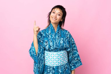 İzole edilmiş mavi arka planda kimono giyen genç bir kadın en iyinin işareti olarak parmağını kaldırıyor.