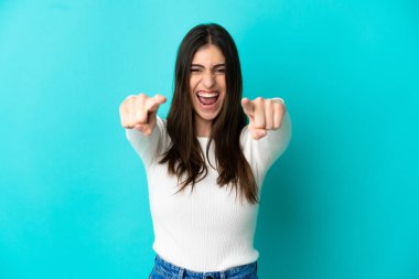 Mavi arka planda izole edilmiş genç beyaz kadın şaşırmış ve ön tarafı işaret ediyor.