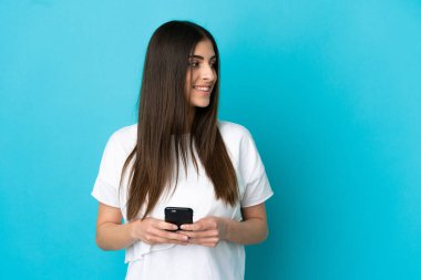 Kafkasyalı genç bir kadın mavi arka planda izole edilmiş ve cep telefonu kullanıyor.