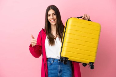 Genç beyaz kadın pembe arka planda izole edilmiş seyahat valiziyle ve başparmağıyla