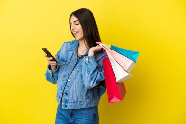 Kafkasyalı genç bir kadın mavi arka planda izole edilmiş. Elinde alışveriş poşetleri var ve bir arkadaşına cep telefonuyla bir mesaj yazıyor.