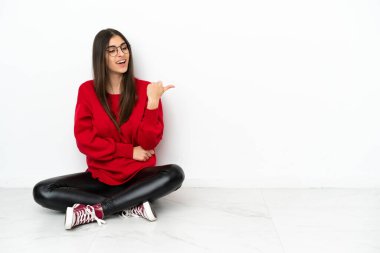 Beyaz arka planda izole bir şekilde yerde oturan genç bir kadın bir ürünü sunmak için kenarı işaret ediyor.