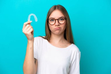 Görünmez diş telleri tutan Litvanyalı genç bir kadın hüzünlü bir ifadeyle mavi arka planda izole edilmiş.