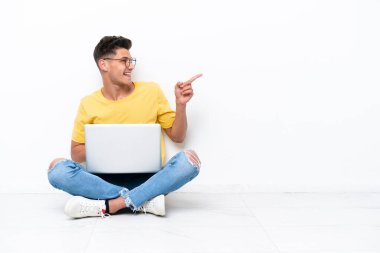 Genç adam, beyaz arka planda izole bir şekilde yerde oturuyor. Parmağını kenarı gösteriyor ve bir ürün sunuyor.