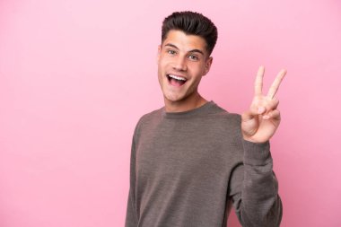 Pembe arka planda izole edilmiş genç beyaz adam gülümsüyor ve zafer işareti gösteriyor.