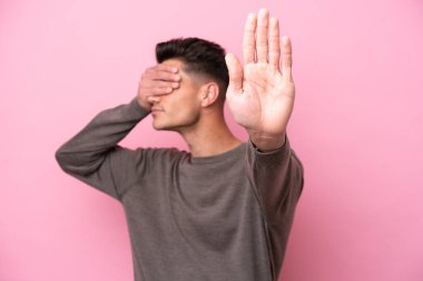 Pembe arka planda izole edilmiş genç beyaz adam jesti durdurup yüzünü kapatıyor.