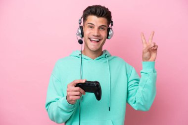 Pembe arka planda izole edilmiş bir bilgisayar oyunu kontrolörüyle oynayan yakışıklı beyaz adam gülümsüyor ve zafer işareti gösteriyor.