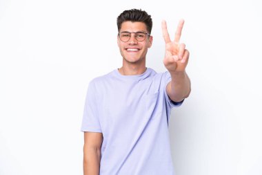 Beyaz arka planda izole edilmiş genç beyaz adam gülümsüyor ve zafer işareti gösteriyor.