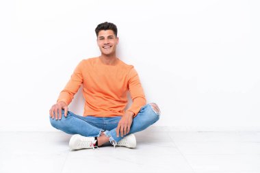 Genç adam beyaz arka planda tek başına yerde oturmuş gülüyor.