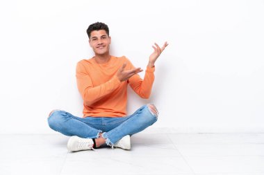 Genç adam beyaz arka planda izole bir şekilde yerde oturuyor. Davet edilmek için ellerini yana uzatıyor.