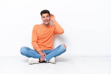 Genç adam beyaz arka planda izole bir şekilde yerde oturuyor. Kulağına elini sürerek bir şeyler dinliyor.