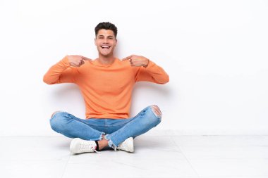 Yerde oturan genç adam beyaz arka planda kendini beğenmiş ve gururlu.