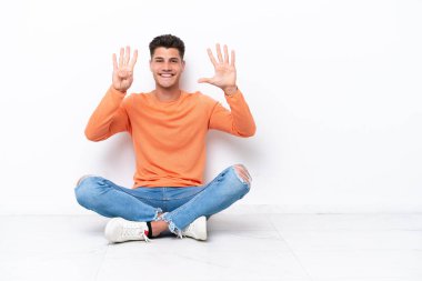 Yerde oturan genç adam beyaz arka planda tecrit edilmiş dokuz parmaklı sayıyor.