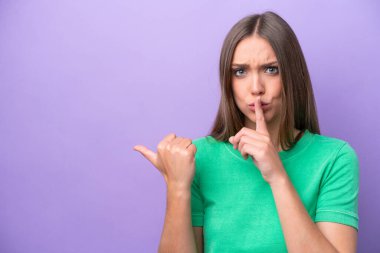 Mor arka planda izole edilmiş genç beyaz bir kadın yan tarafı işaret ediyor ve sessiz hareketler yapıyor.