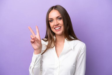 Mor arka planda izole edilmiş genç beyaz kadın gülümsüyor ve zafer işareti gösteriyor.