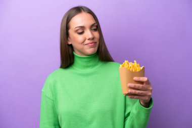 Mor arka planda kızarmış patates kızartması tutan genç beyaz kadın yana bakıyor ve gülümsüyor.