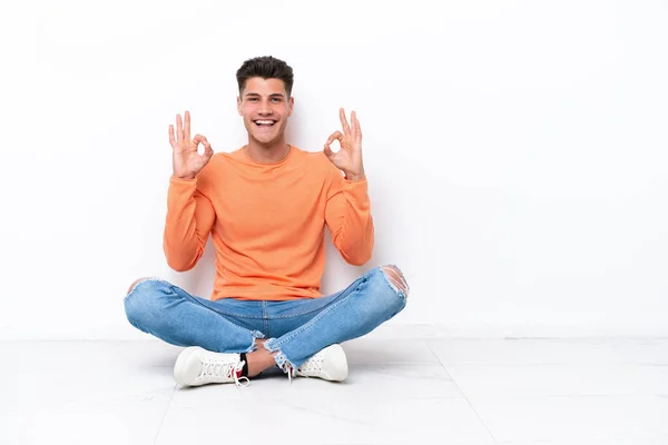 Mladý Muž Sedící Podlaze Izolovaný Bílém Pozadí Ukazující Znamení Prsty — Stock fotografie