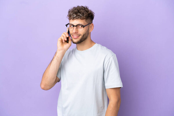 Delivery caucasian man isolated on purple background keeping a conversation with the mobile phone with someone
