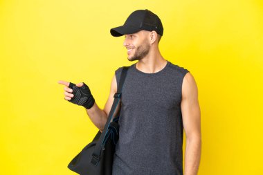 Sarı arka planda izole edilmiş spor çantalı genç sarışın bir adam. Parmakla göstererek bir ürün sunuyor.