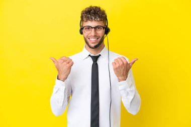 Sarı arka planda izole edilmiş bir kulaklıkla çalışan pazarlamacı sarışın adam. Baş parmakları havada ve gülümsüyor.