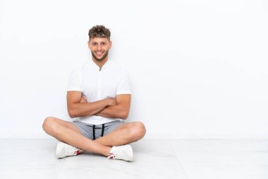 Genç sarışın adam, beyaz arka planda izole bir şekilde yerde oturuyor ve kollarını ön cephede kavuşturuyor.