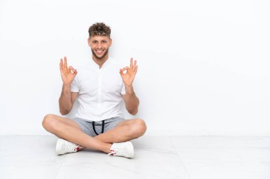 Beyaz arka planda izole bir şekilde yerde oturan genç sarışın adam parmaklarıyla 