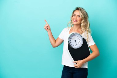 Mavi arka planda izole edilmiş, tartı makinesi ve işaret eden tarafı olan genç Brezilyalı kadın.