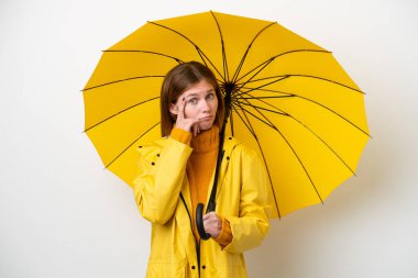 Yağmura dayanıklı ceketi ve şemsiyesi olan genç bir İngiliz kadın beyaz arka planda izole edilmiş bir fikir düşünüyor.