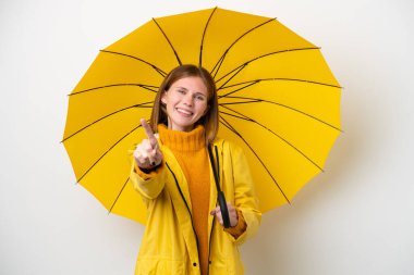 Yağmura dayanıklı ceketi ve şemsiyesi olan genç bir İngiliz kadını beyaz arka planda izole edilmiş ve parmağını kaldırıyor.