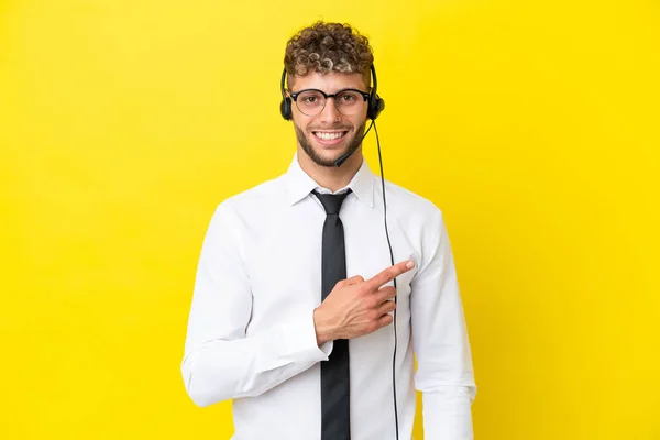 一位金发碧眼的电话推销员 带着一只黄色背景的耳机 一边指着侧面 一边展示自己的产品 — 图库照片