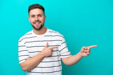 Mavi arka planda izole edilmiş genç beyaz adam şaşırmış ve işaret ediyor.