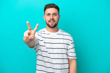 Mavi arka planda izole edilmiş genç beyaz adam gülümsüyor ve zafer işareti gösteriyor.