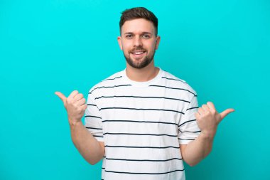 Kafkasyalı genç adam mavi arka planda izole edilmiş. Baş parmakları havada ve gülümsüyor.