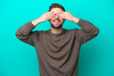 Mavi arka planda izole edilmiş genç beyaz bir adam elleriyle gözlerini kapatıyor ve gülümsüyor.