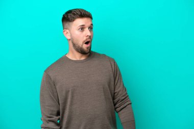 Mavi arka planda izole edilmiş genç beyaz adam yan tarafa bakarken sürpriz jest yapıyor.