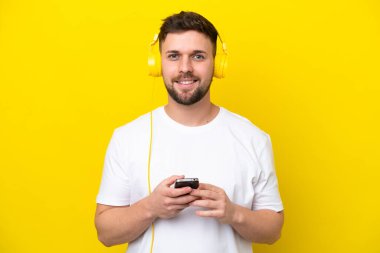 Kafkasyalı genç adam sarı arka planda izole edilmiş. Cep telefonuyla müzik dinliyor ve ön tarafa bakıyor.