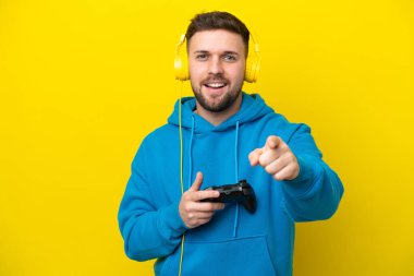 Sarı arka planda izole edilmiş bir bilgisayar oyunu kontrolörüyle oynayan genç beyaz adam şaşırmış ve ön tarafı işaret ediyor.